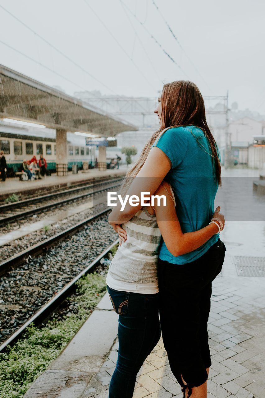 Happy young female friends embracing at railroad station during monsoon