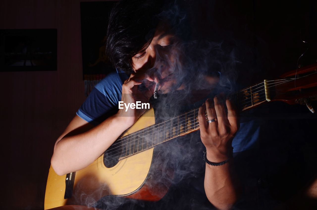 Man smoking cigarette while playing guitar in darkroom