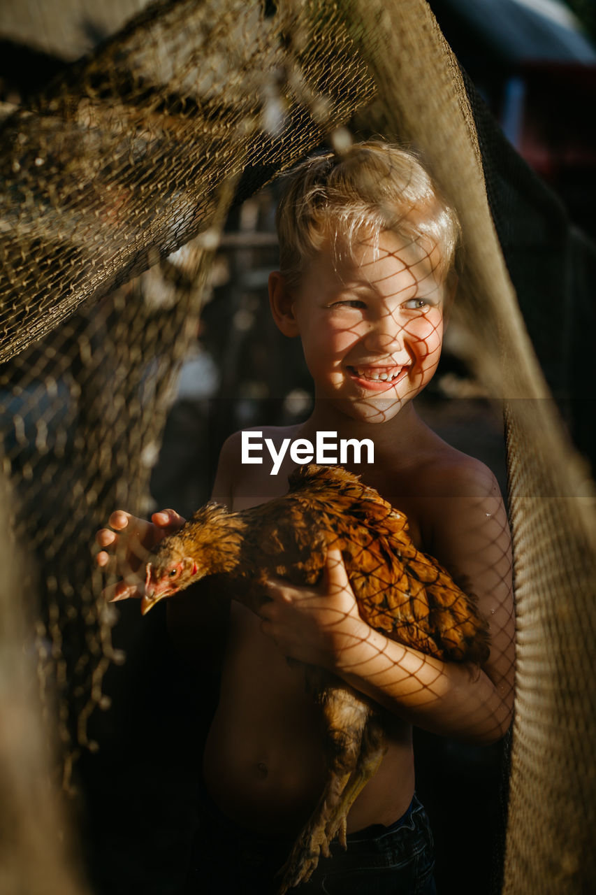 Smiling shirtless boy holding chicken while standing outdoors