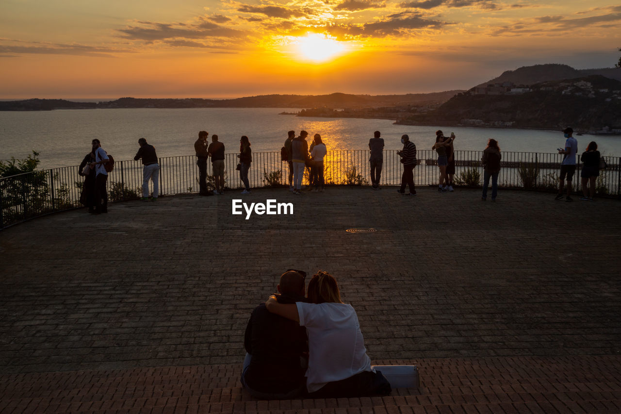 REAR VIEW OF PEOPLE BY SEA AGAINST SUNSET SKY