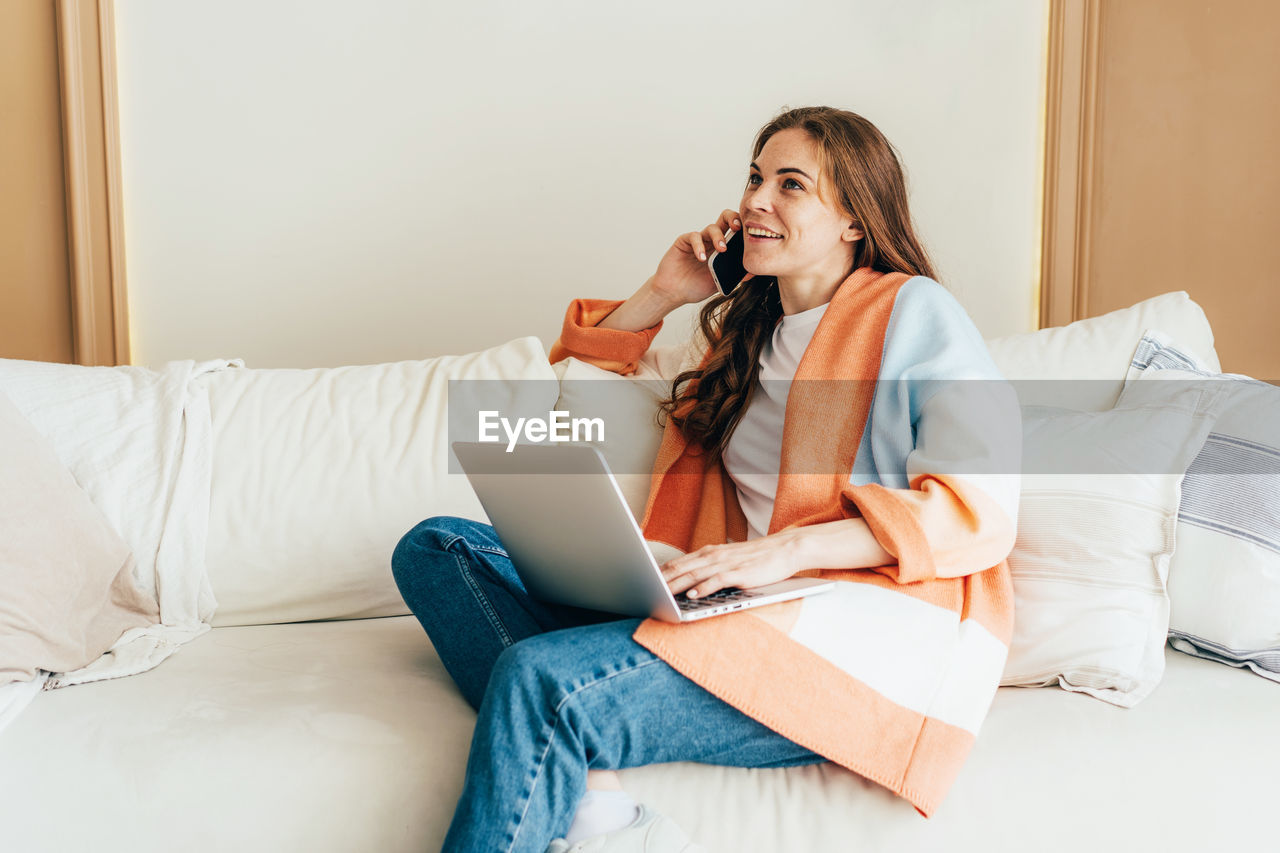Young business woman smiling and talking on phone while work with laptop at home sitting on sofa.