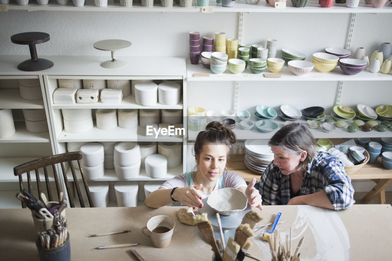 High angle view of mature female potter looking at young craftsperson molding clay at workshop