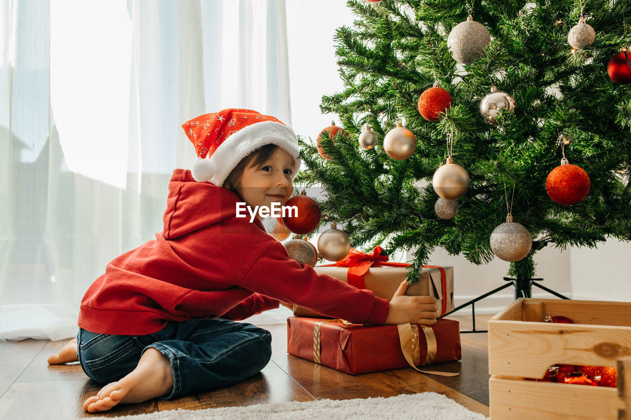 Charming boy in a santa hat takes out christmas gifts from under the new year tree and laughs