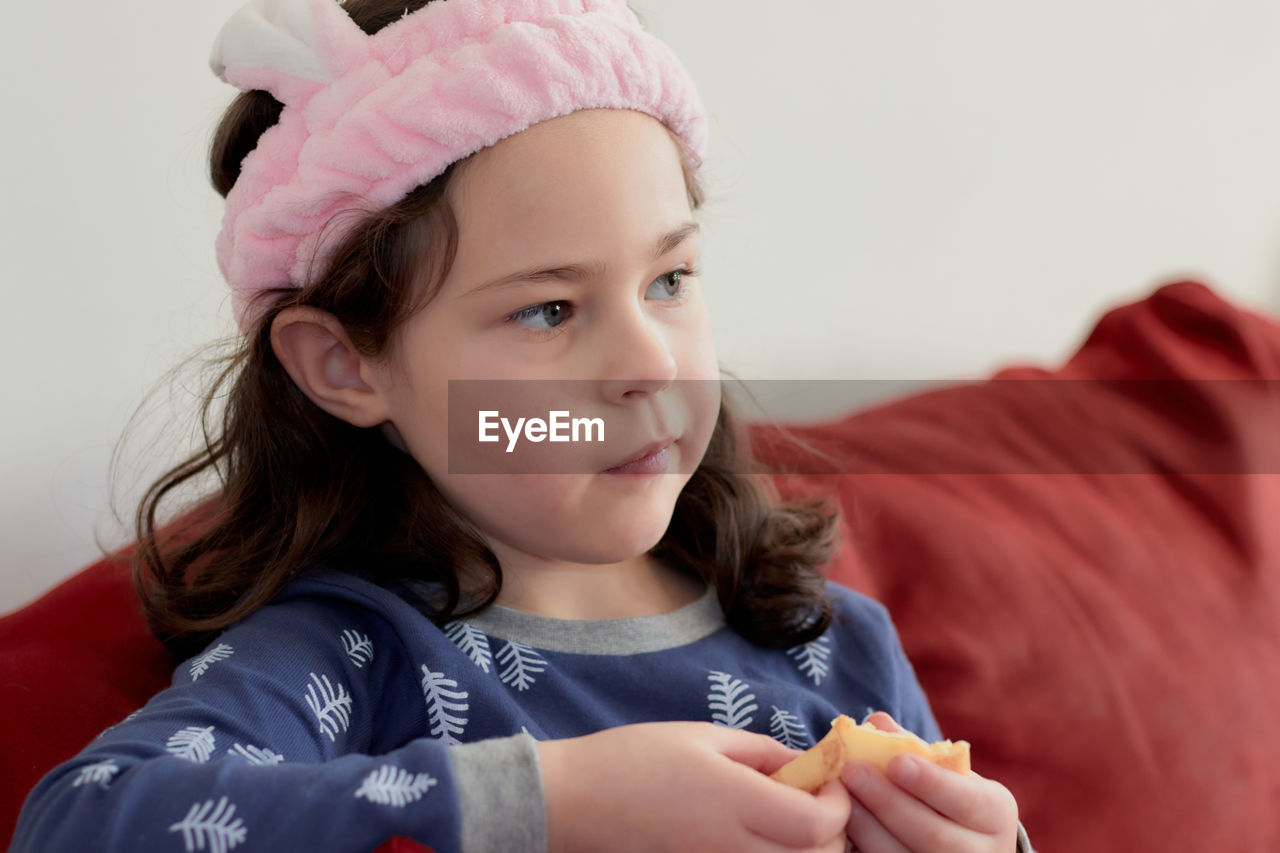 Cute little girl in a pink head dress is eating crepes on the couch