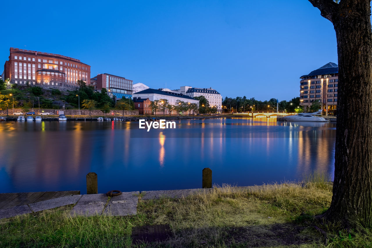 Illuminated buildings by lake against blue sky at dusk