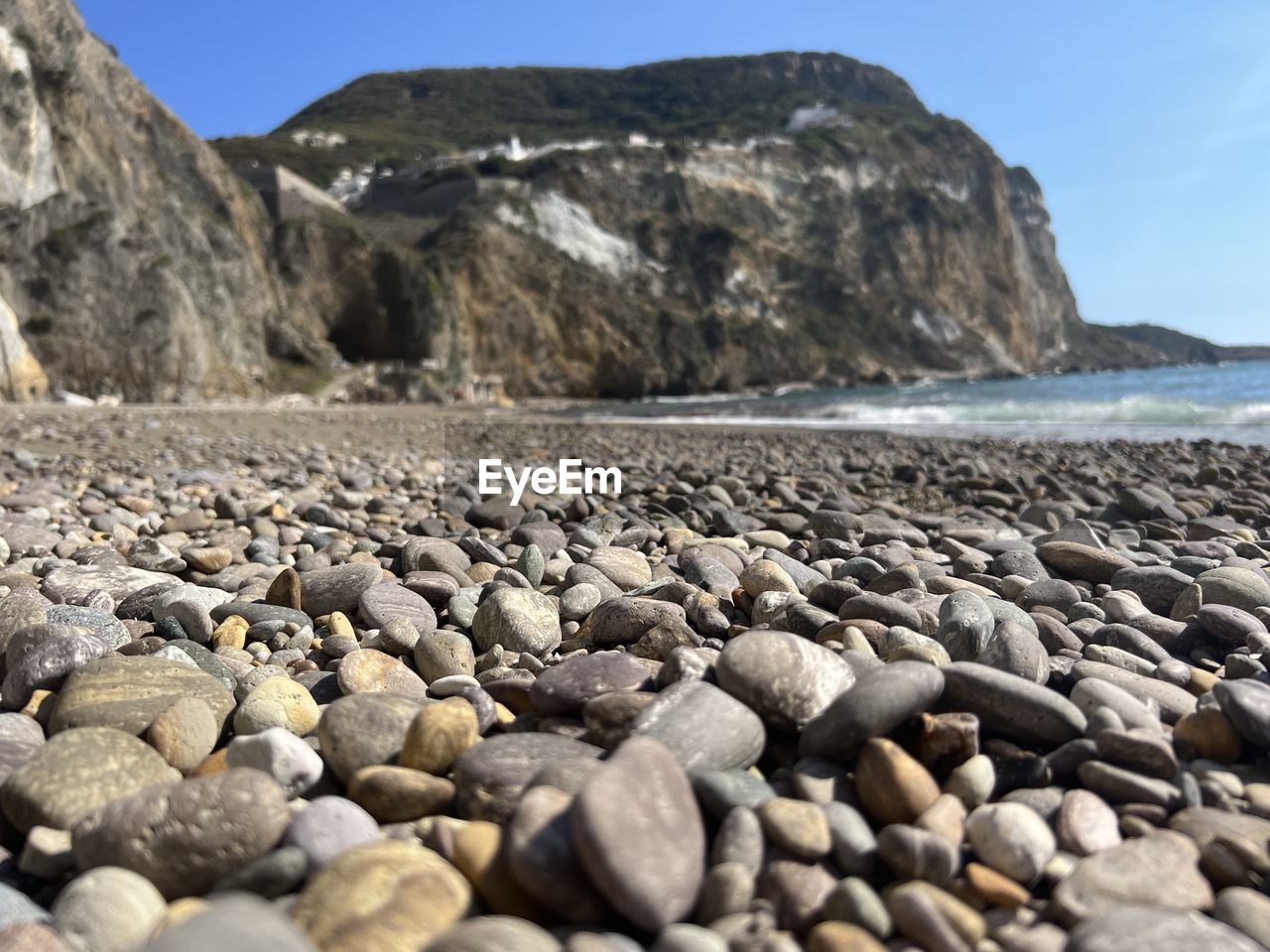 rock, land, water, sea, beach, sky, nature, shore, coast, scenics - nature, beauty in nature, pebble, stone, no people, tranquility, geology, mountain, environment, tranquil scene, landscape, day, travel destinations, coastline, outdoors, travel, blue, clear sky, water's edge, gravel, sand, coastal feature, non-urban scene, sunny, sunlight