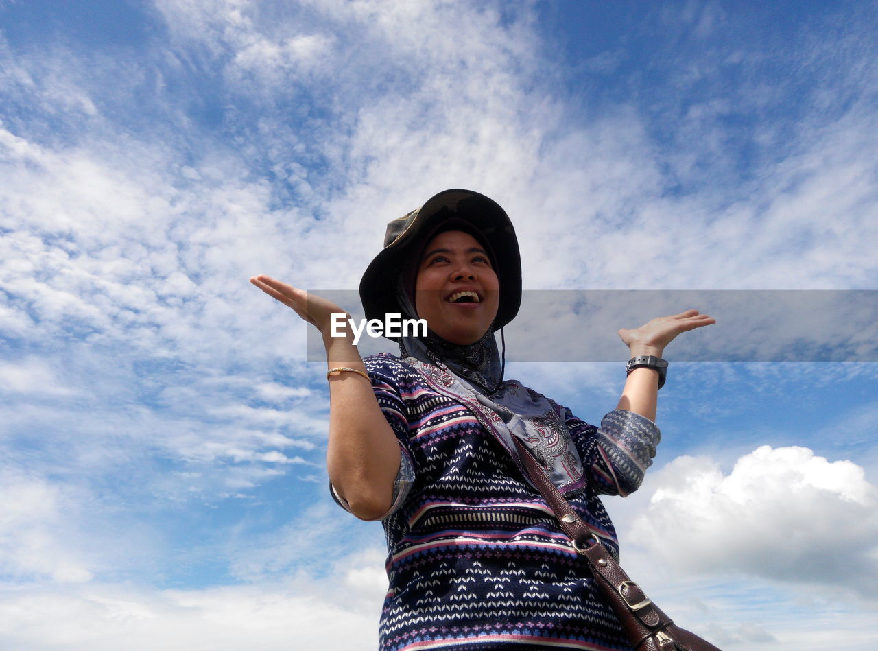 Low angle view of woman with hands raised laughing against cloudy sky