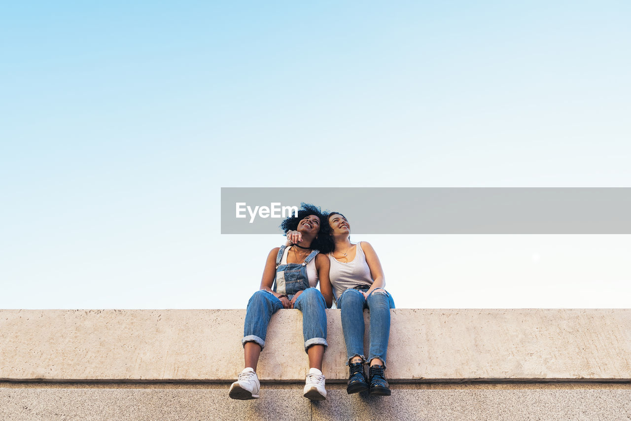 Low angle view of female friends taking selfie while sitting on retaining wall