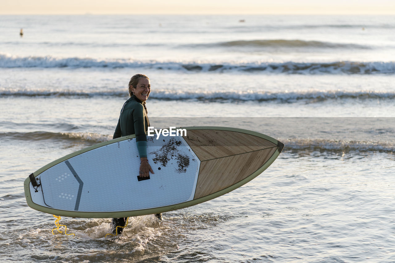 Woman laughing while holding paddleboard standing in sea during dawn