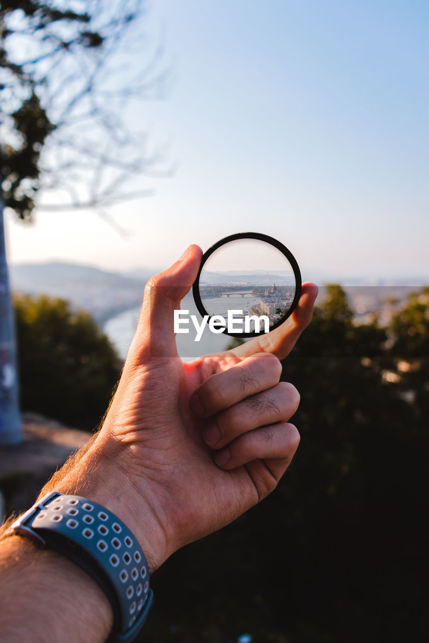 Close-up of hand holding magnifying glass against sky