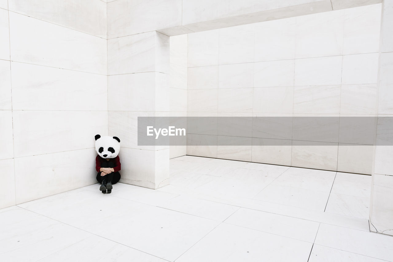 Sad woman wearing panda mask sitting in corner against wall