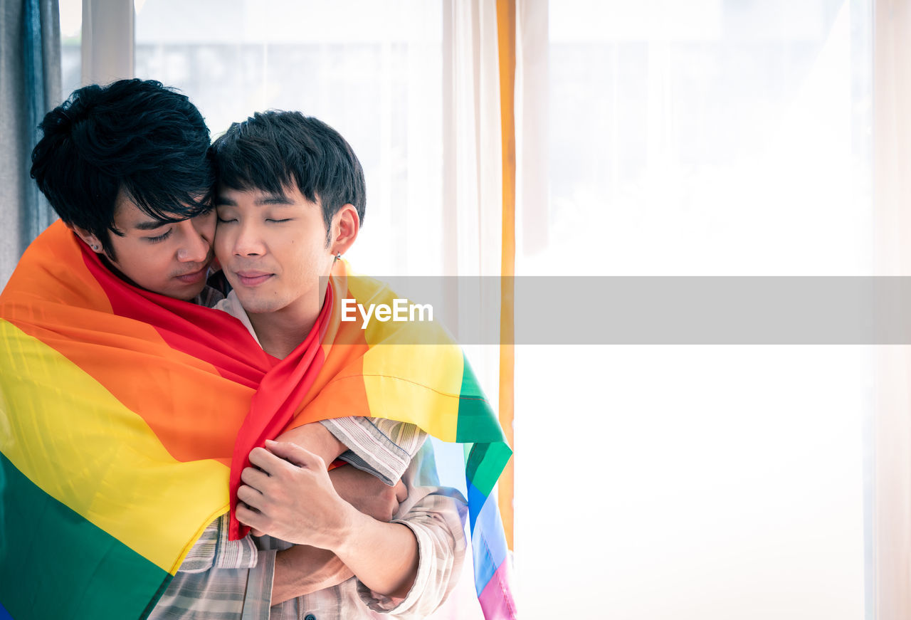 Gay couple holding rainbow flag while standing against window at home