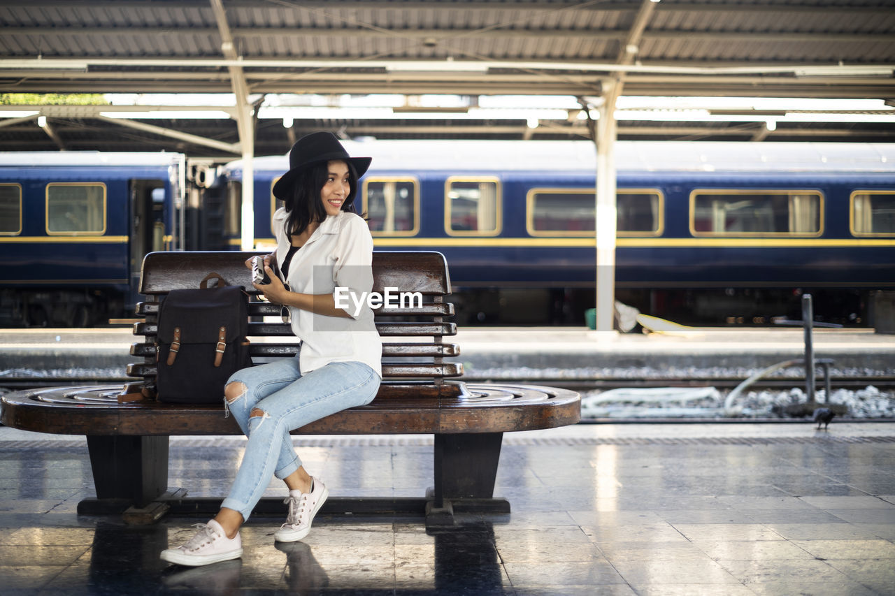 Full length of woman holding camera sitting at railroad station platform