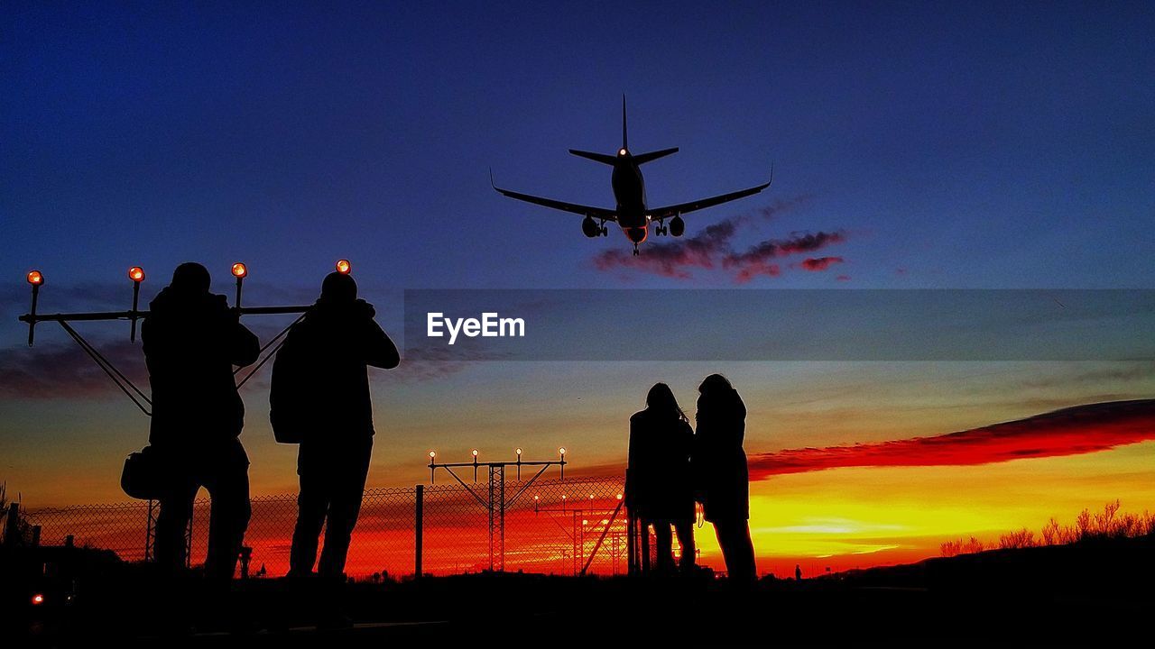 Silhouette people standing against orange sky with airplane