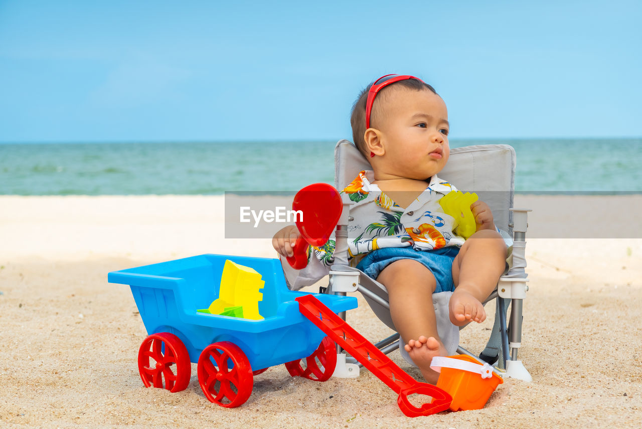 CUTE BOY SITTING ON BEACH WITH TOY ON SHORE