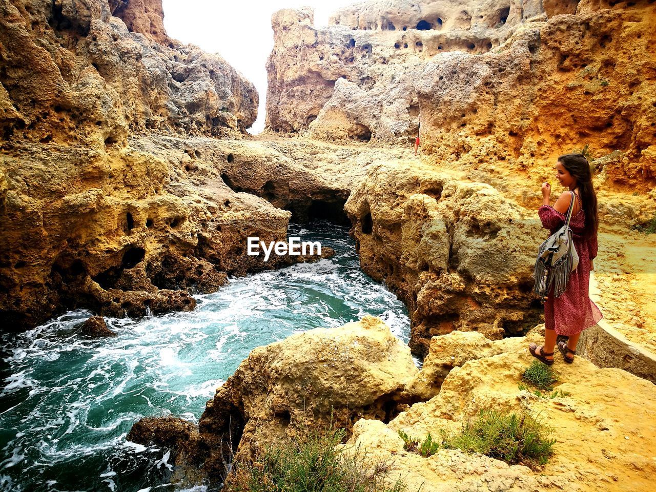 REAR VIEW OF WOMAN PHOTOGRAPHING BY ROCK FORMATION IN WATER