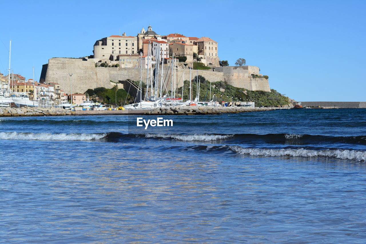 Buildings by sea against clear blue sky