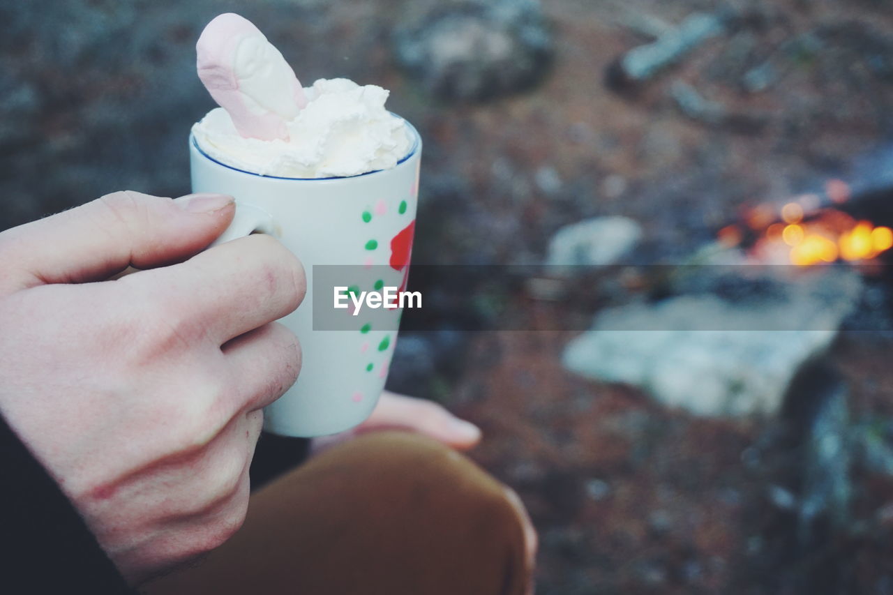 Cropped image of man holding hot chocolate by campfire in forest