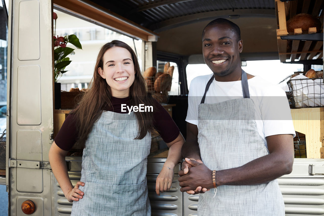 Portrait of smiling male and female owners standing outside food truck parked in city