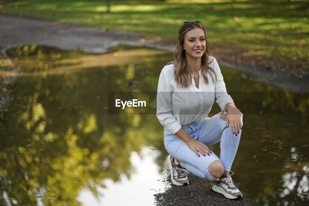 Portrait of smiling young woman crouching by water at park