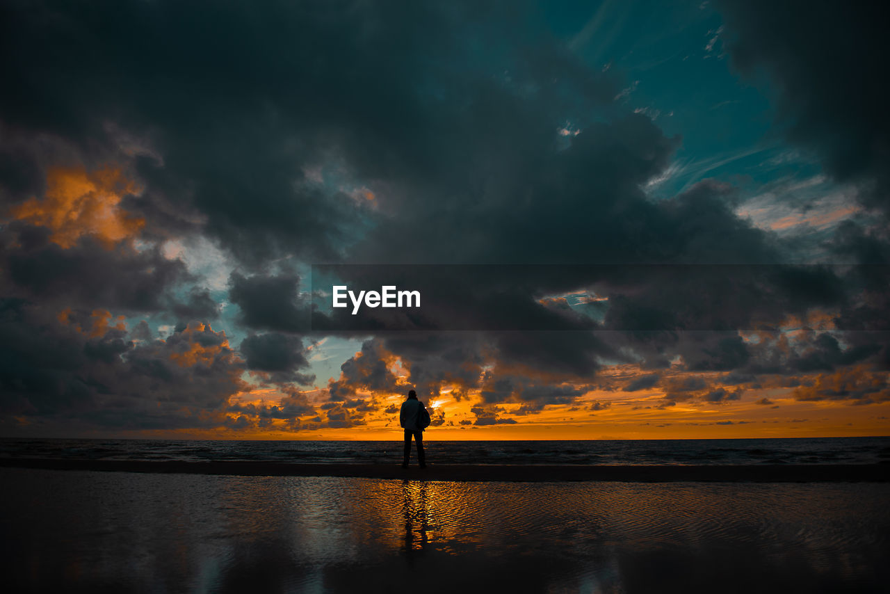 Silhouette man standing at beach against cloudy sky during sunset