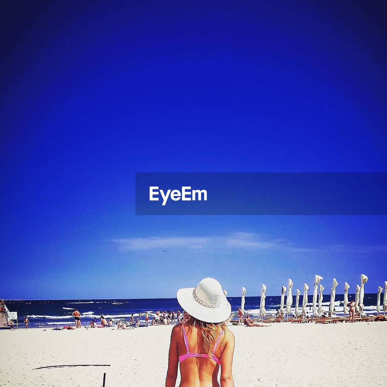 Rear view of woman in bikini wearing sun hat at beach against blue sky