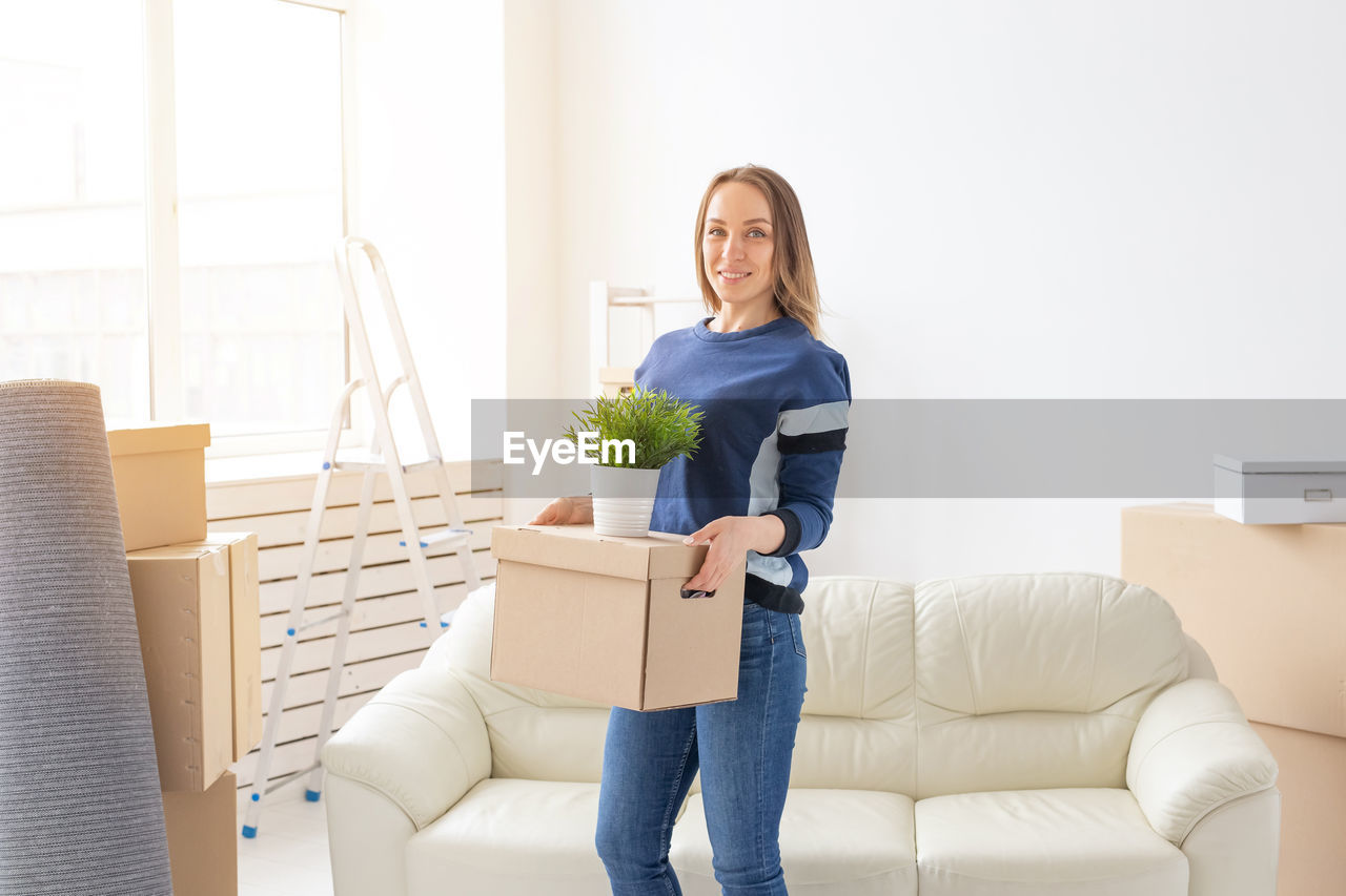 PORTRAIT OF SMILING YOUNG WOMAN HOLDING SOFA IN BOX AT HOME