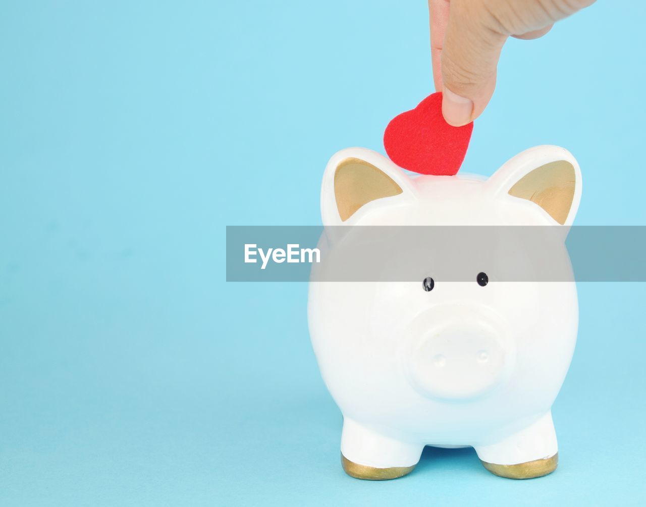 Person hand putting heart shape paper in piggy bank against blue background