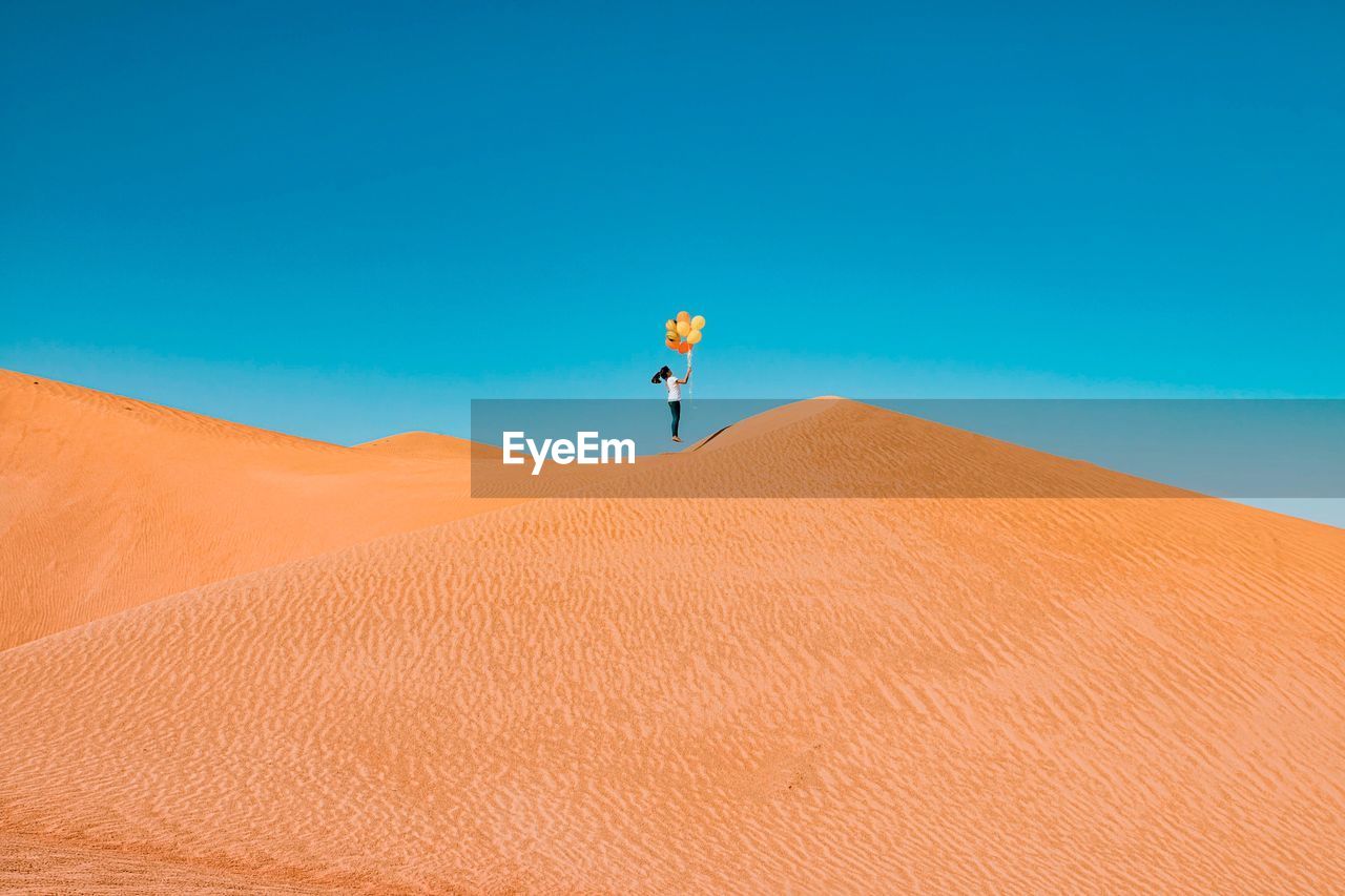 Low angle view of girl holding balloons while jumping on sand dune at desert