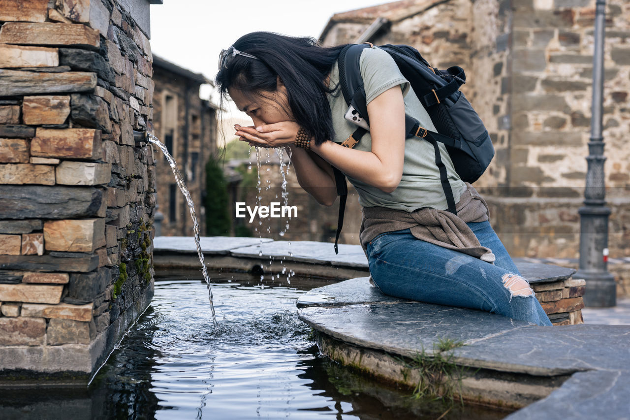 Asian female tourist with backpack drinking water from fountain on street with aged stone buildings in old town during trip in valverde de los arroyos