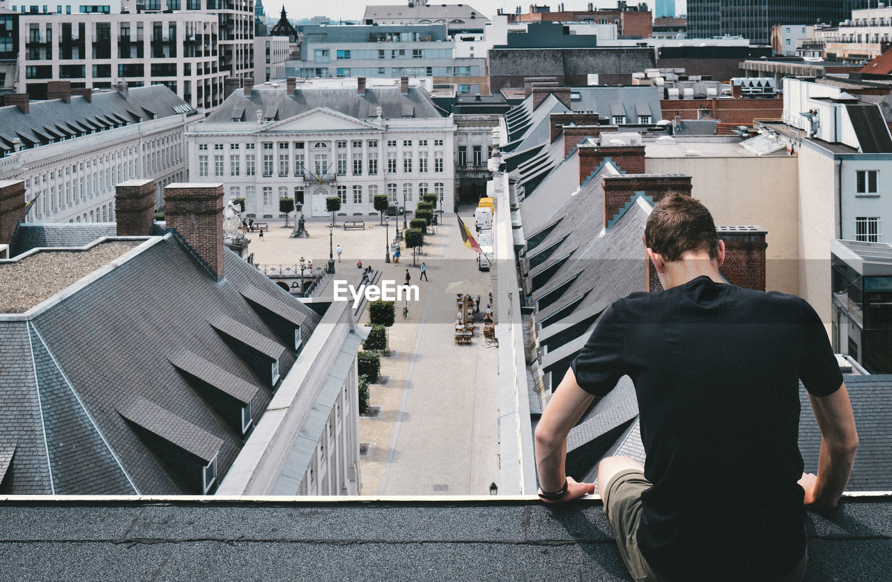 Rear view of man sitting on retaining wall in city