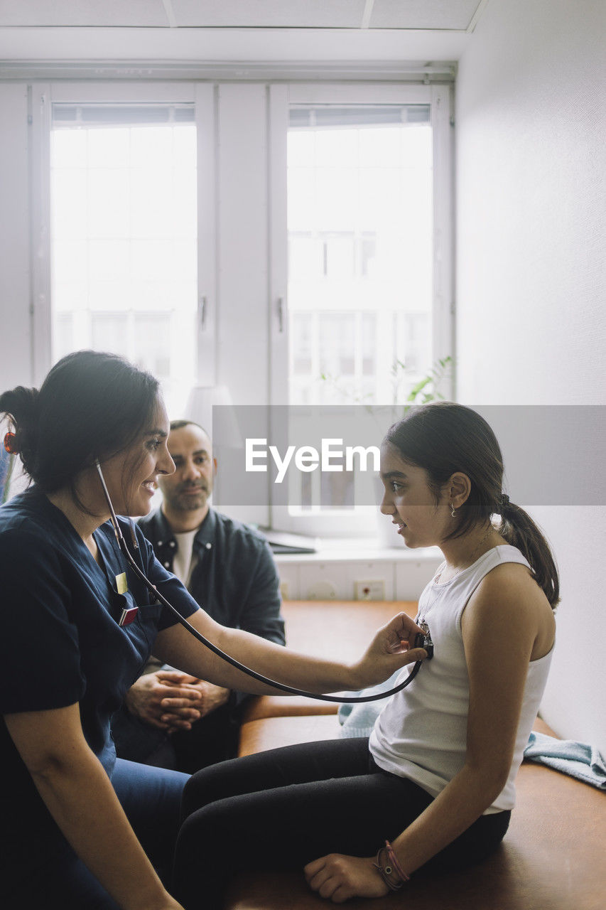 Smiling female nurse listening to heartbeat of patient sitting on bed at hospital