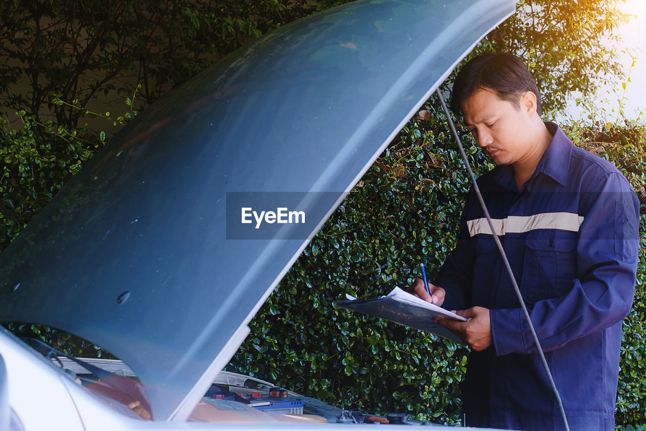 Mechanic writing in notepad by car against plants