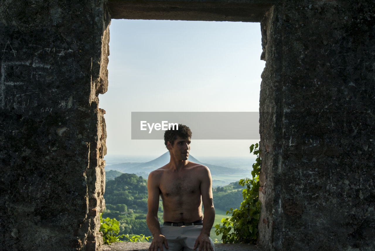 Shirtless man sitting on window sill against sky