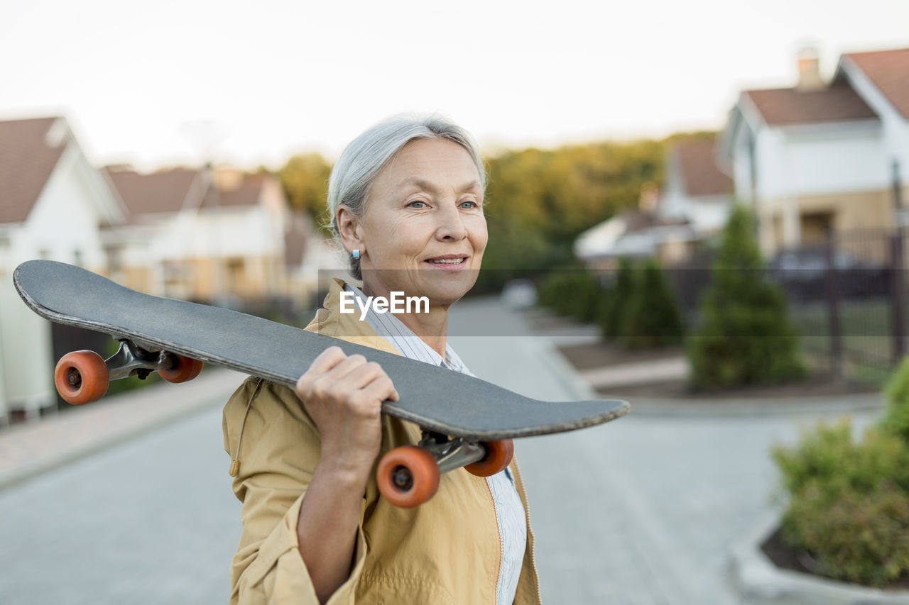 Portrait of smiling senior woman with skateboard on her shoulder