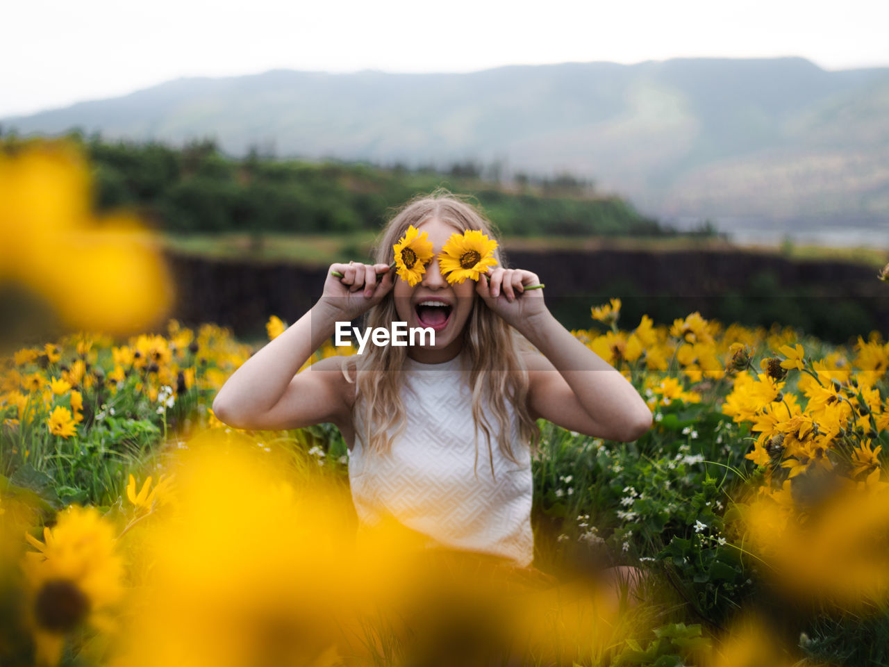 Excited woman shouting while holding flowers over eyes by plants against sky