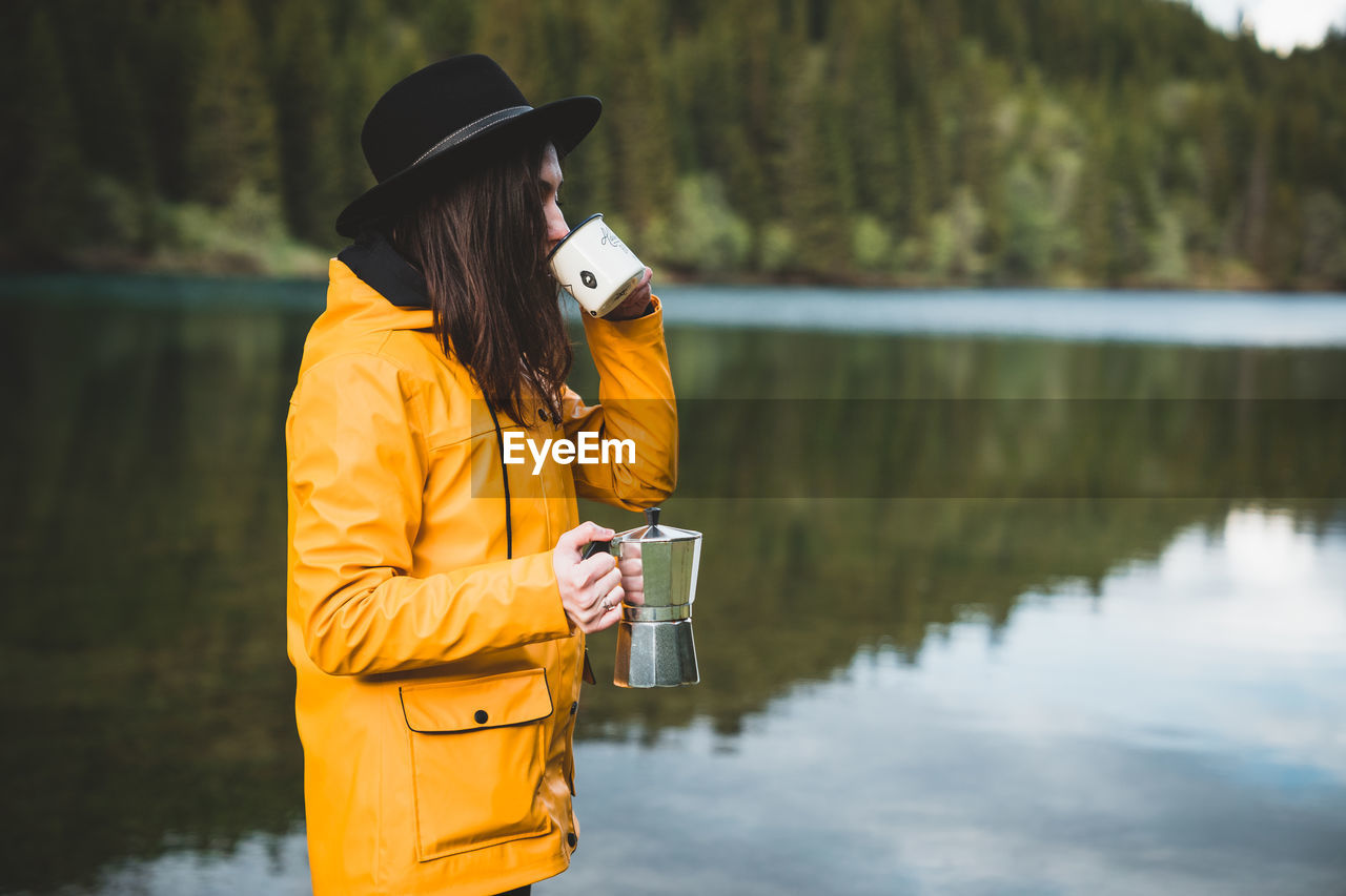 Side view of stylish female hipster wearing a hat, holding and drinking coffee from moka by the lake