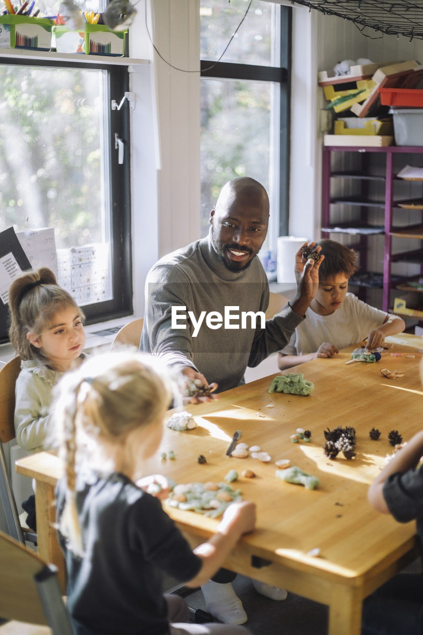 Male teacher playing with kids while sitting at bench during art class at preschool