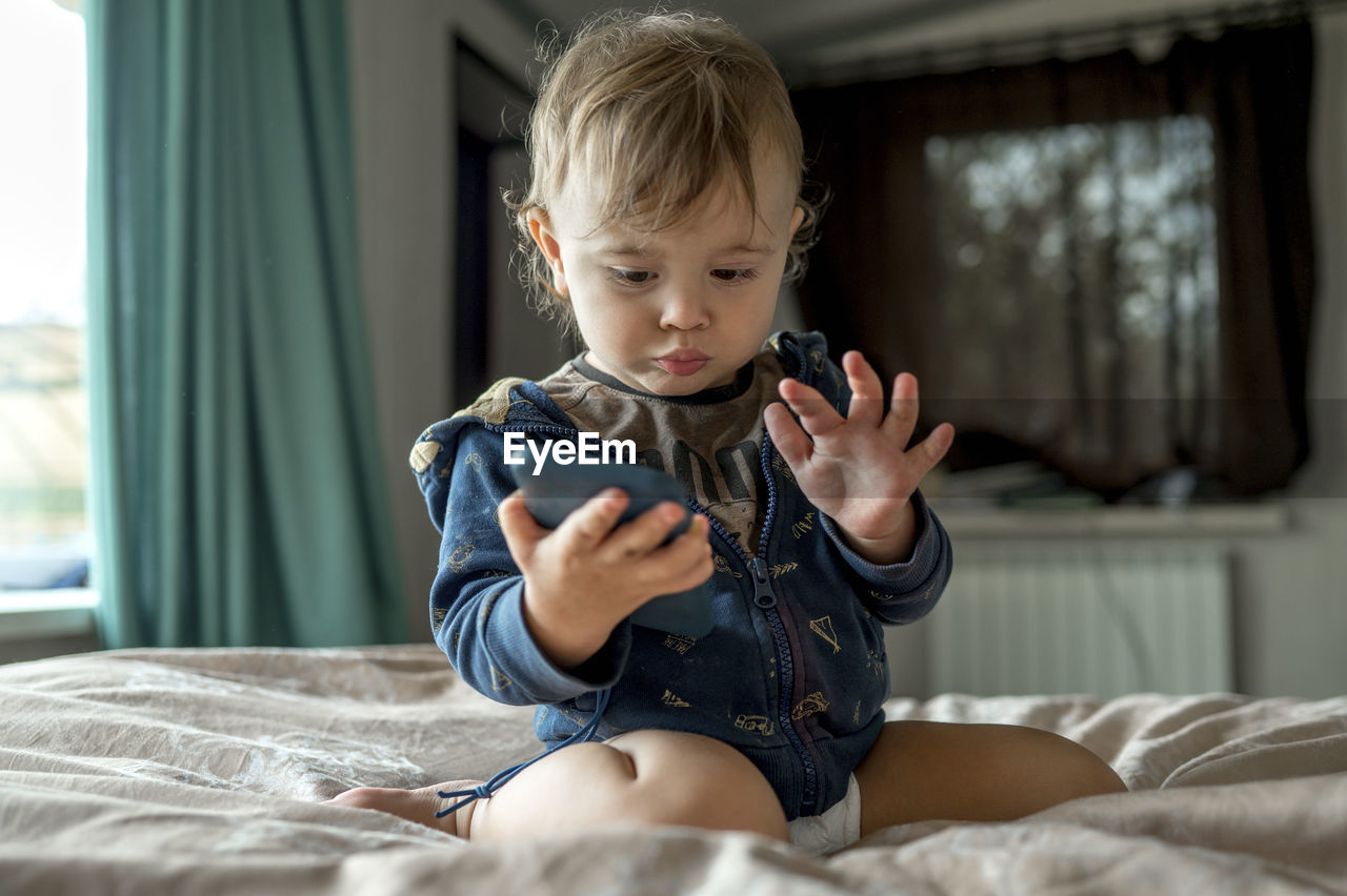 Cute baby boy looking at mobile phone on bed