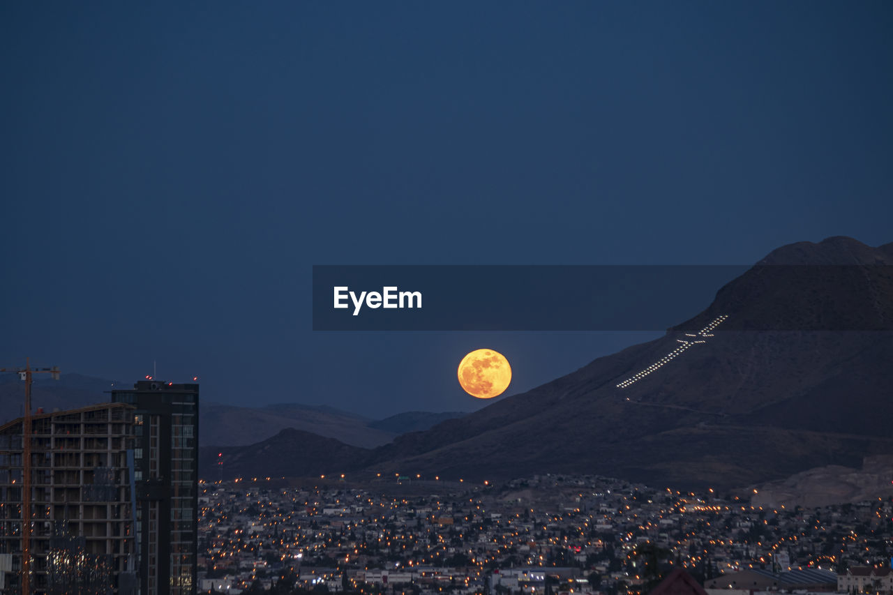 Aerial view of moon over illuminated city against clear sky at dusk