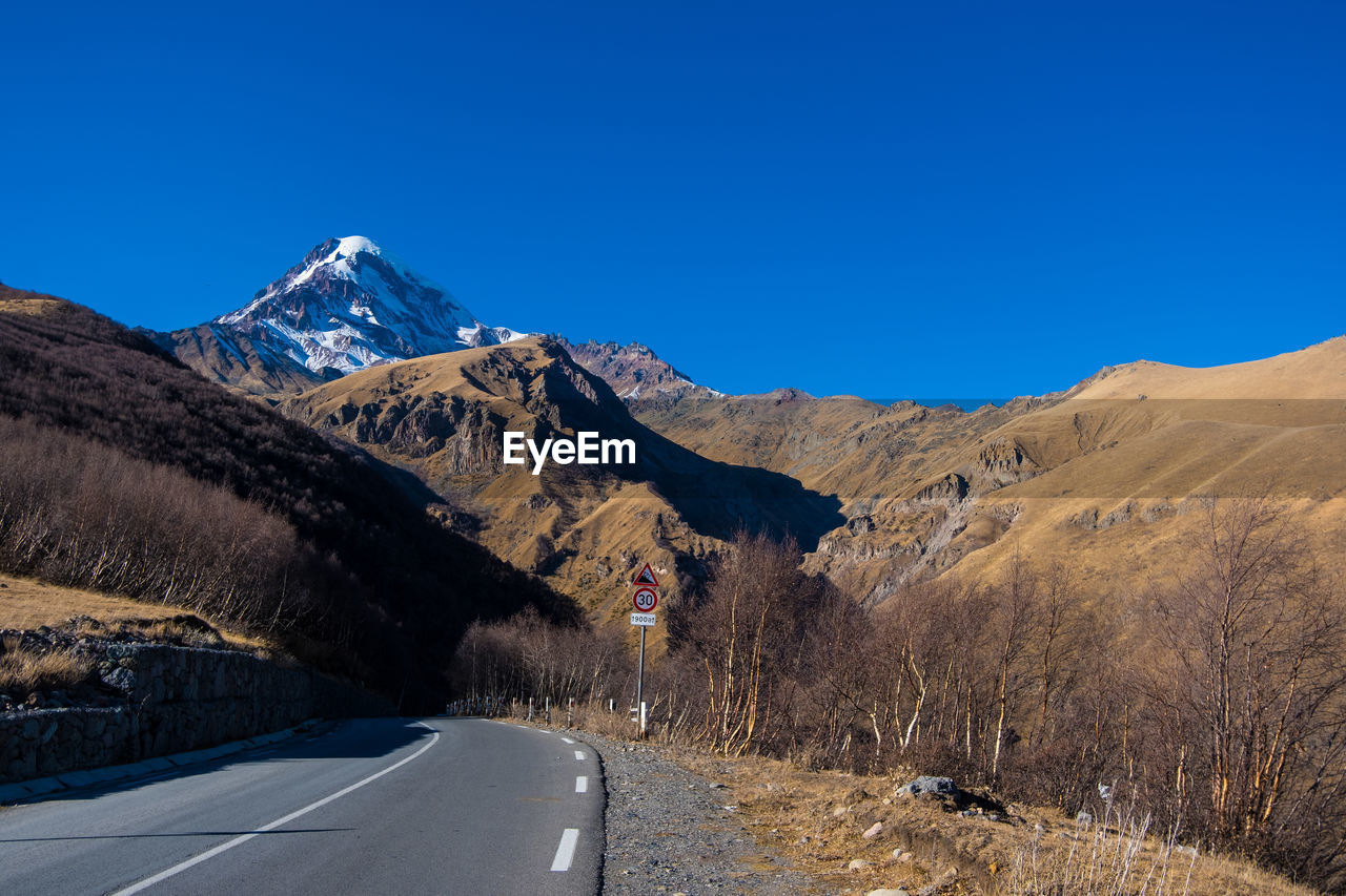 mountain, road, sky, scenics - nature, mountain range, landscape, environment, transportation, mountain pass, nature, clear sky, beauty in nature, blue, snow, no people, travel, the way forward, travel destinations, winter, land, cold temperature, non-urban scene, sunny, valley, tranquil scene, highway, plant, tranquility, day, outdoors, tree, snowcapped mountain, ridge, symbol, empty road, remote, sunlight, infrastructure, tourism, plateau, country road, wilderness, mountain peak