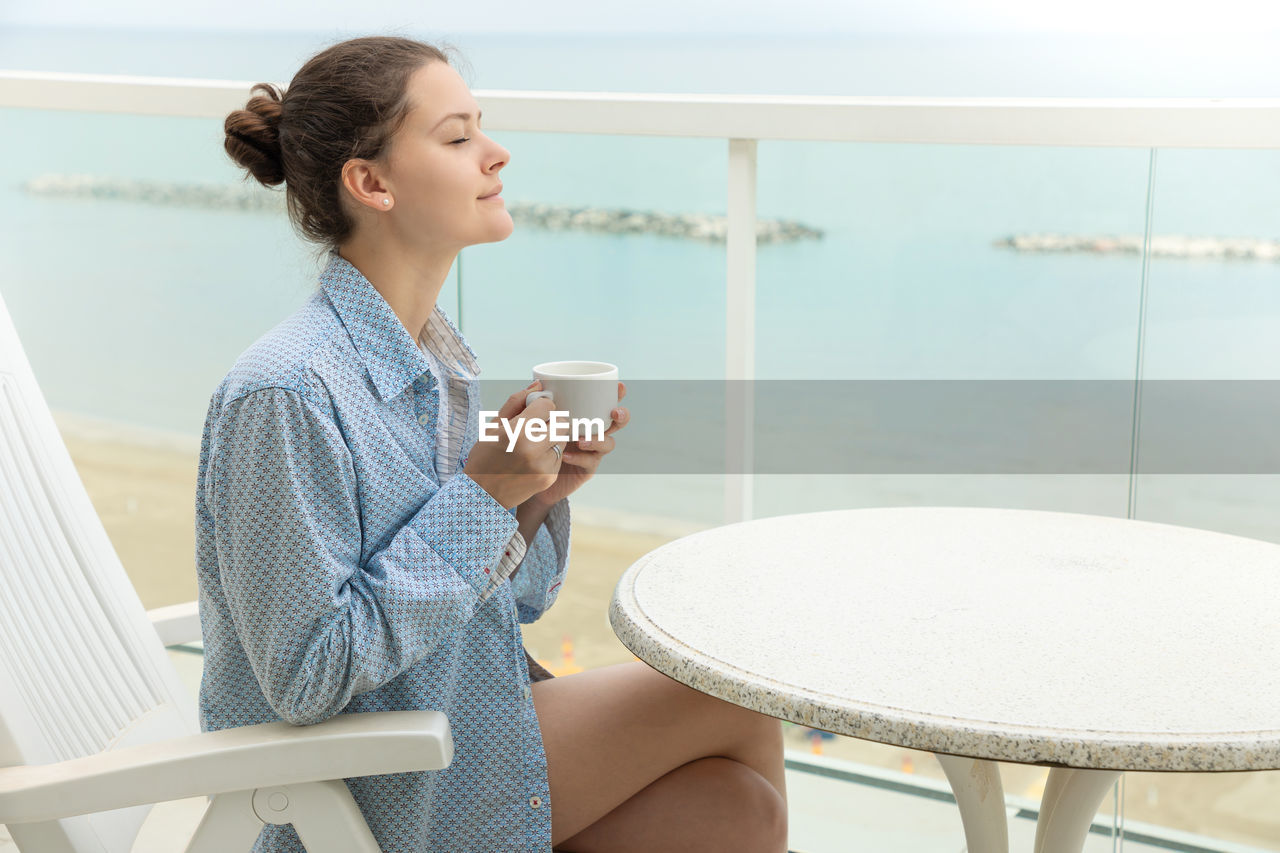 YOUNG WOMAN DRINKING COFFEE WHILE SITTING ON CHAIR AT TABLE