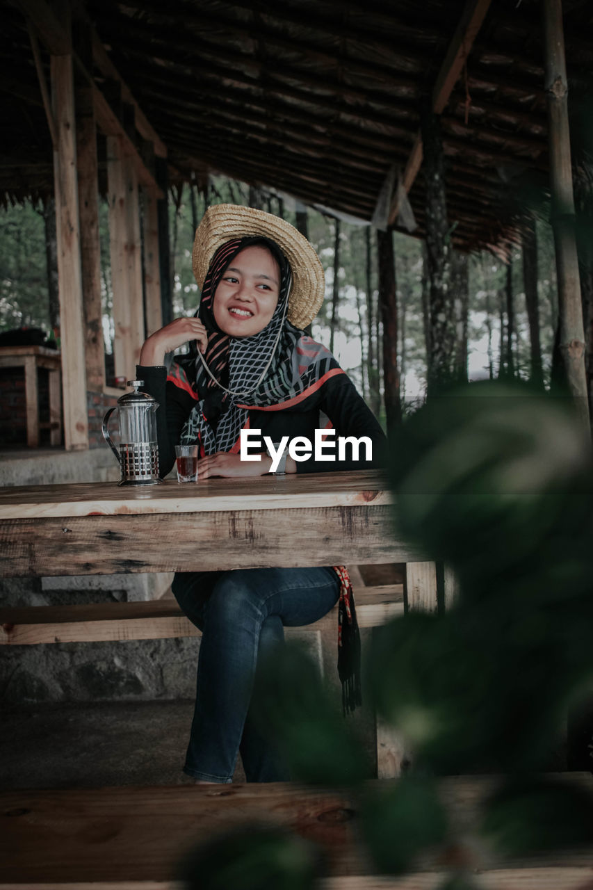 Smiling young woman wearing hat looking away while sitting at table