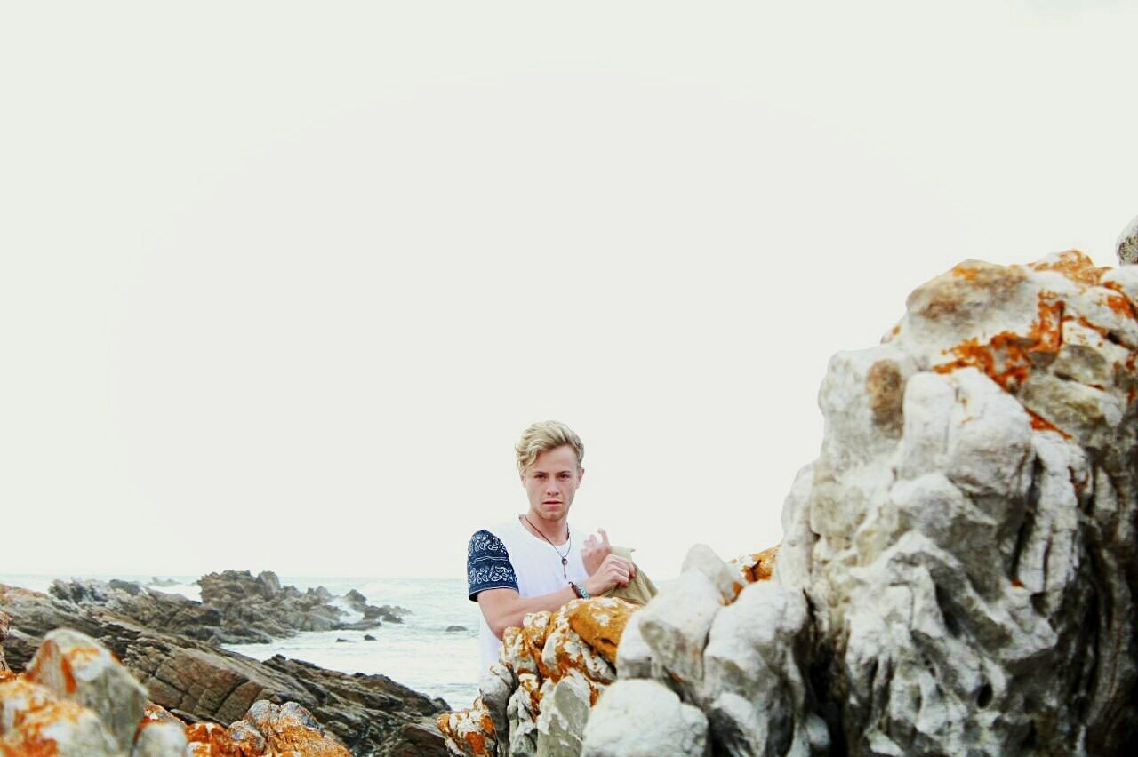 Portrait of young man standing by rocks on shore against clear sky