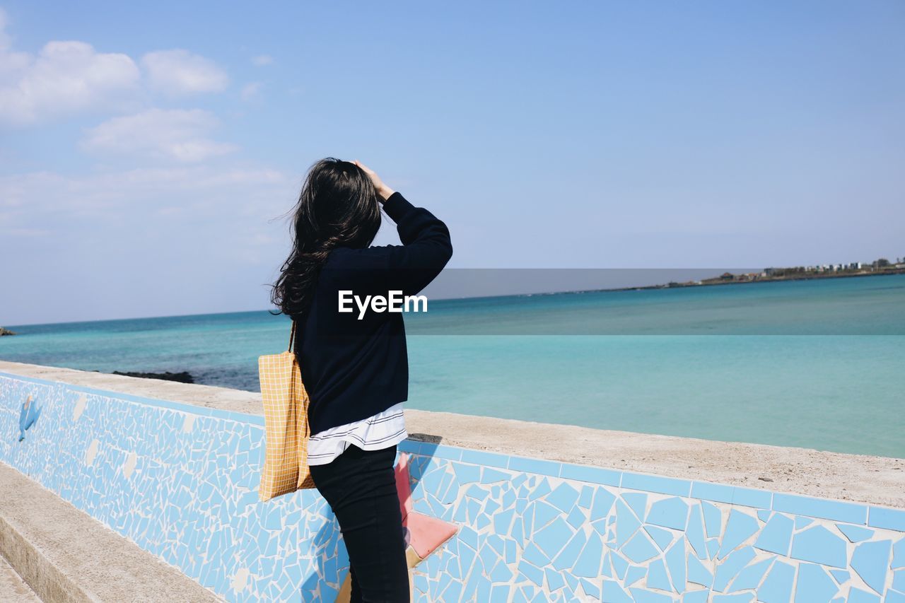 Side view of woman looking at sea while standing by retaining wall against blue sky during sunny day