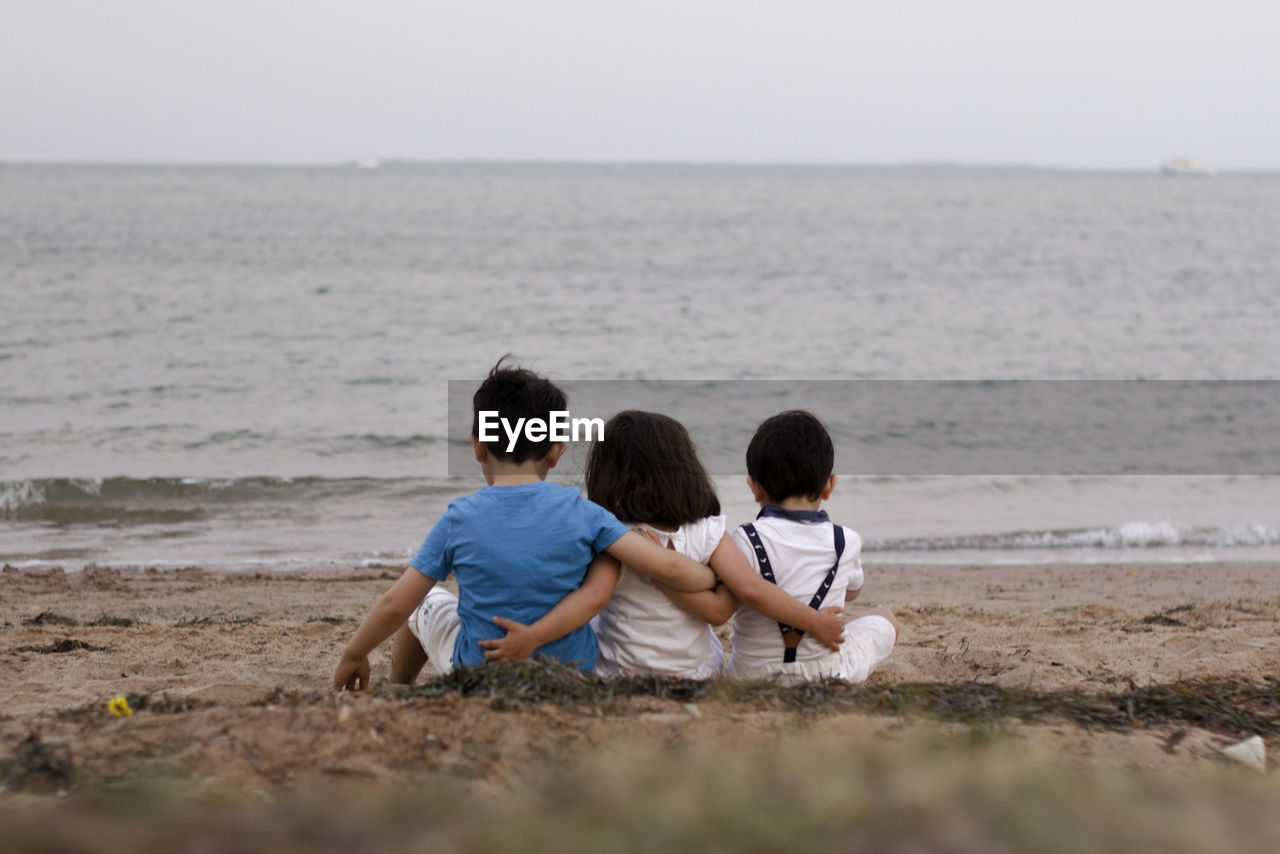 Three kids setting down on the sand while hugging each other