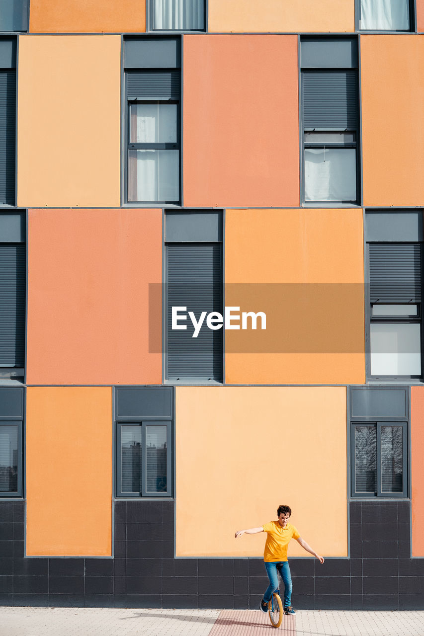 Full body of young guy in casual wear riding on unicycle on modern urban street with colorful building