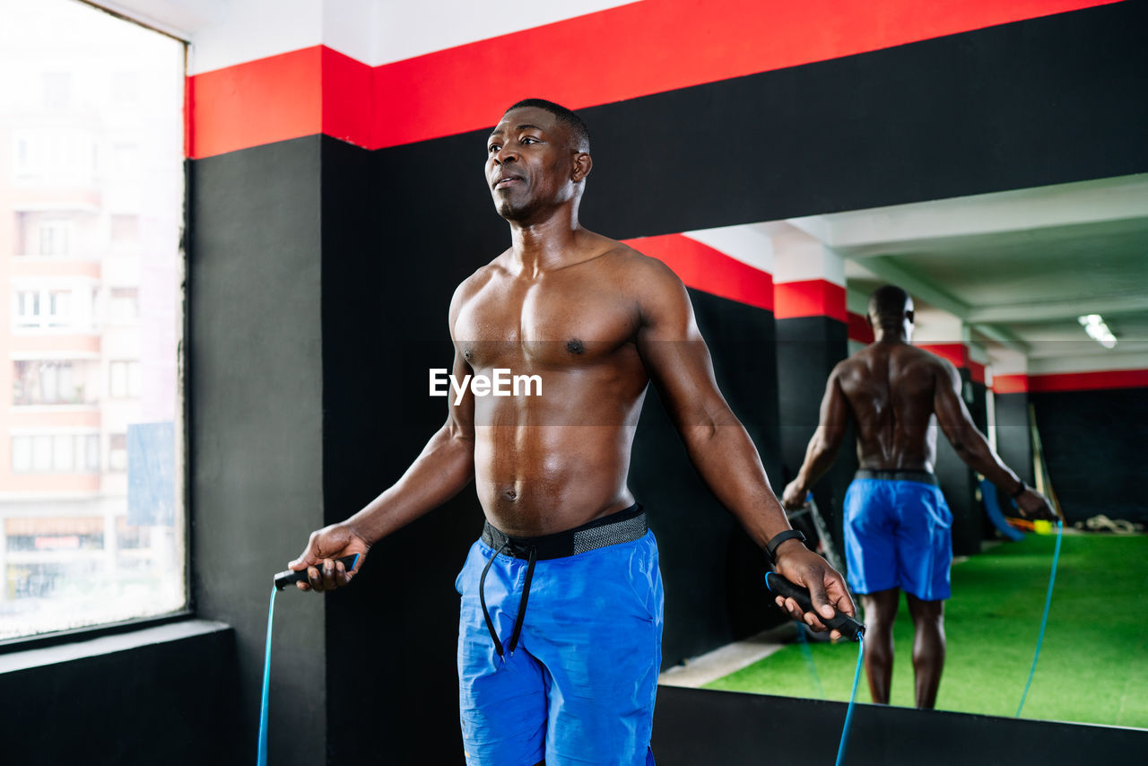 Sweaty african american sportsman with muscular body skipping rope in modern bright fitness center during functional workout