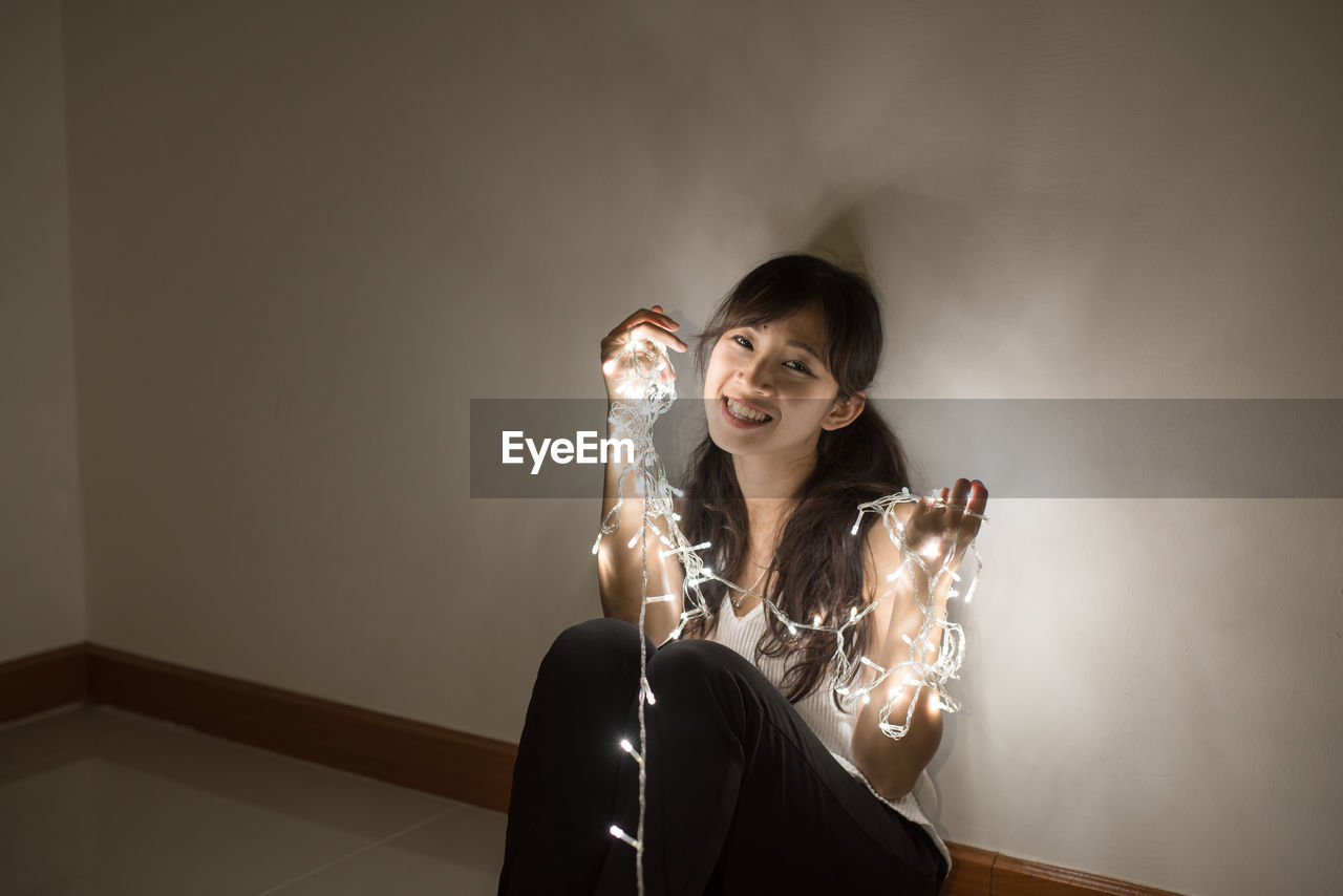 Young woman with illuminated string light sitting by wall at home