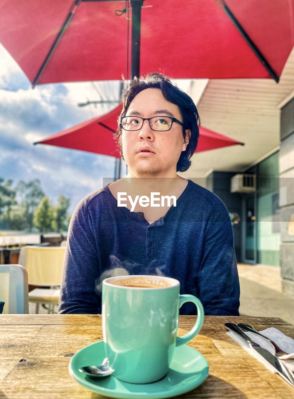 Portrait of young asian man having hot coffee at sidewalk cafe under red cafe umbrella.